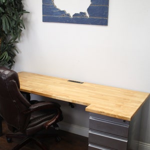 May include: A light wood desk with a gray metal file cabinet and a brown leather office chair. The desk has a cutout for cords and a blue wooden wall art with a white cutout of the United States.