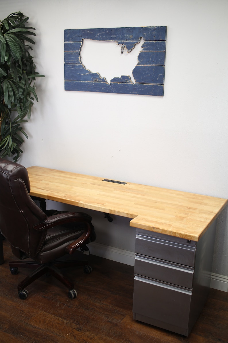 May include: A light wood desk with a gray metal file cabinet and a brown leather office chair. The desk has a cutout for cords and a blue wooden wall art with a white cutout of the United States.