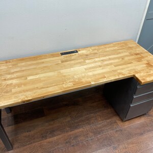 May include: A wooden desk with a light wood top and black metal legs. The desk has a built-in power strip and a set of drawers on the right side.