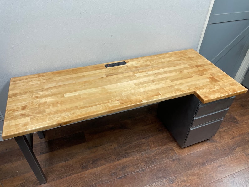 May include: A wooden desk with a light wood top and black metal legs. The desk has a built-in power strip and a set of drawers on the right side.