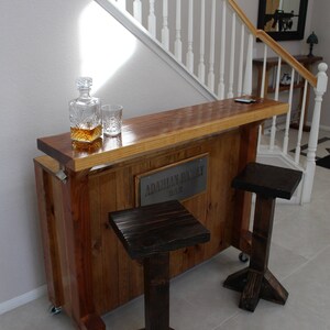 May include: A wooden bar with a sign that reads "Adamian Family Bar" and two dark brown wooden stools. The bar is made of reclaimed wood and has a rustic look.