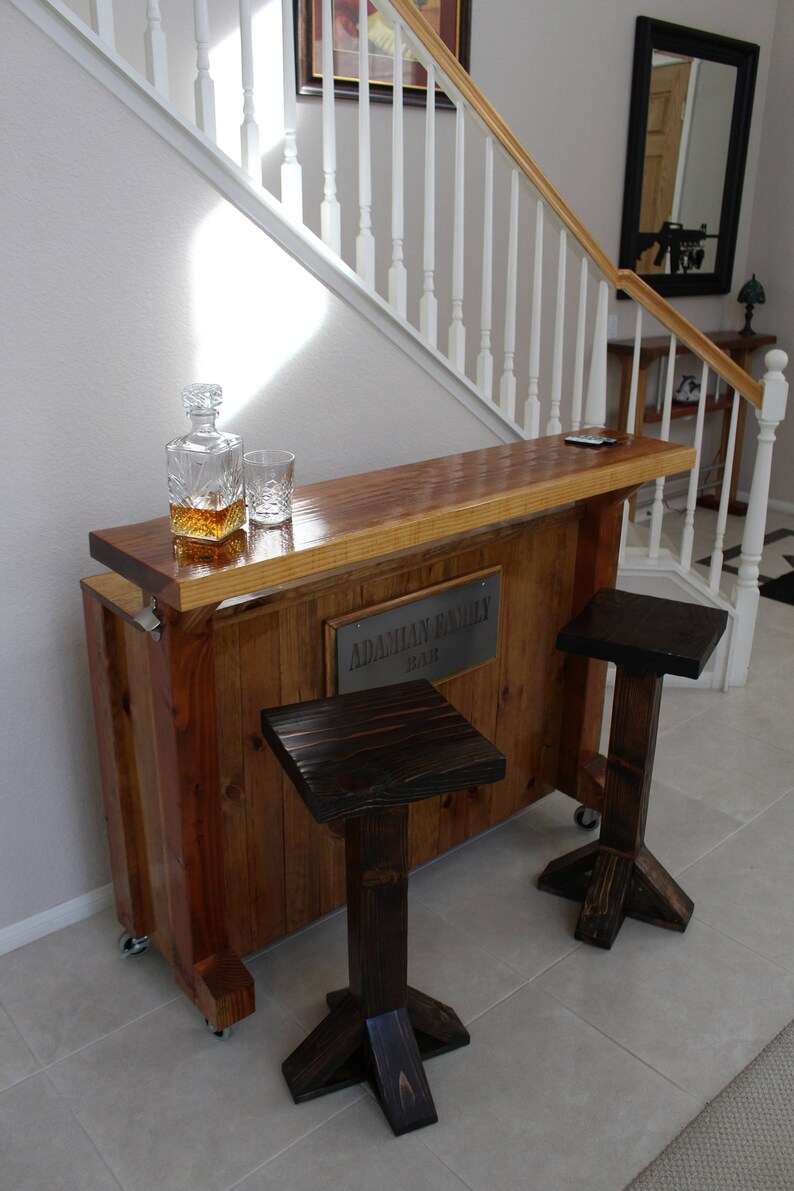 May include: A wooden bar with a sign that reads "Adamian Family Bar" and two dark brown wooden stools. The bar is made of reclaimed wood and has a rustic look.