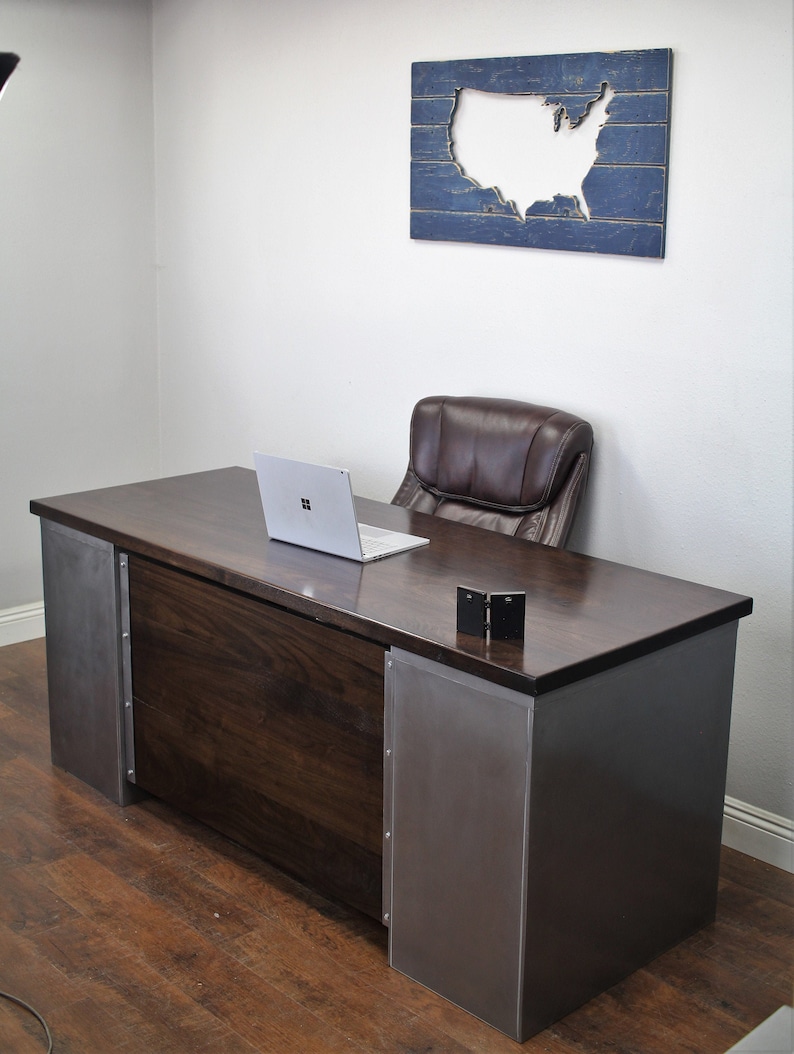 May include: A dark wood desk with metal accents. The desk has a laptop computer on the surface and a chair behind it. A wooden sign with a cutout of the United States is on the wall behind the desk.
