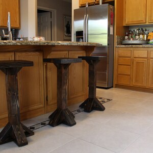 May include: Three dark brown wooden bar stools with a simple design. The stools are in a kitchen with wood cabinets and a stainless steel refrigerator.