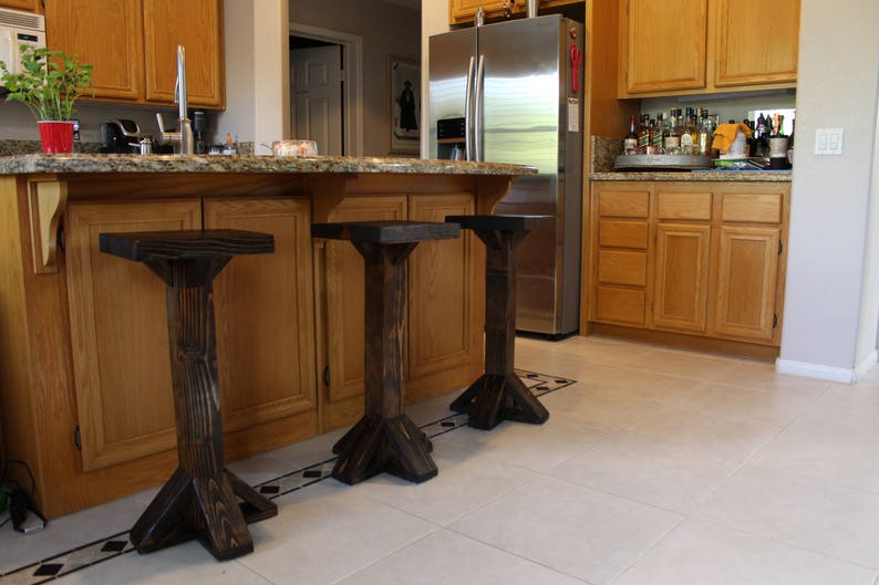 May include: Three dark brown wooden bar stools with a simple design. The stools are in a kitchen with wood cabinets and a stainless steel refrigerator.