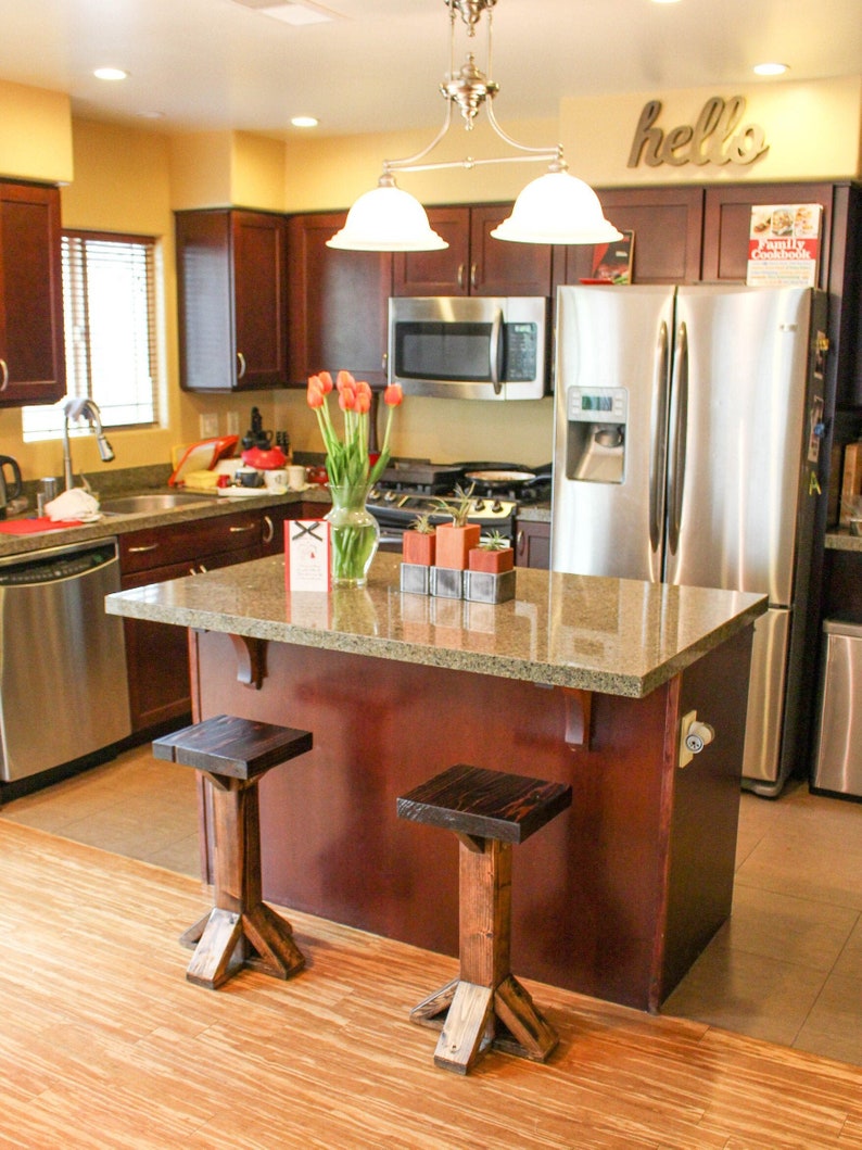 May include: A kitchen island with a granite countertop and a dark wood base. Two wooden stools are positioned in front of the island. The island is located in a kitchen with stainless steel appliances and dark wood cabinets. The word "hello" is written on the wall above the cabinets.
