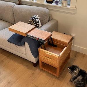 Pode incluir: Uma mesa de apoio de madeira com tampo elevável e duas gavetas. A mesa é feita de madeira de cerejeira e tem um acabamento natural. A mesa está aberta e o tampo está levantado, revelando o interior da mesa. Dois gatos estão na sala, um está sentado no chão e o outro está caminhando em direção à mesa.