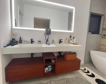 May include: A double vanity with a white countertop and two integrated sinks, featuring a large rectangular LED-backlit mirror above. The base consists of reddish-brown wooden cabinetry with two drawers and a central open shelving unit for storage.