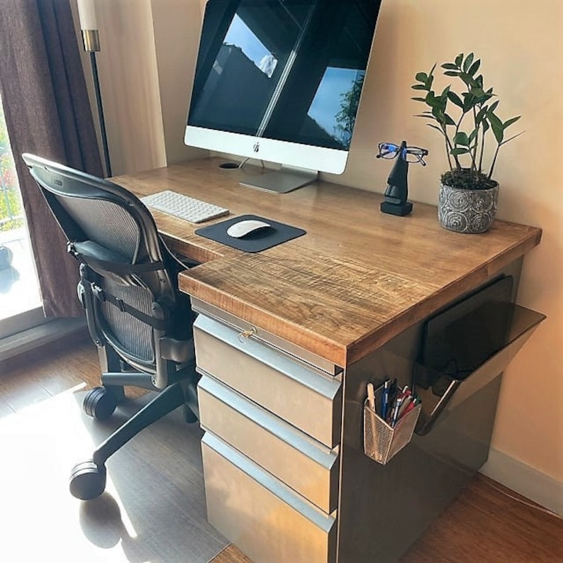 May include: A wooden desk with a metal filing cabinet and a computer monitor. The desk has a light wood finish and the filing cabinet has a brushed metal finish. There is a black office chair with a mesh back and a potted plant on the desk.