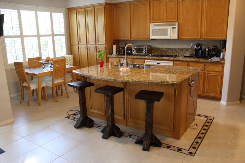 May include: A kitchen island with a granite countertop and three black wooden bar stools. The island has a light wood base and is situated in a kitchen with light wood cabinets and a tiled floor.