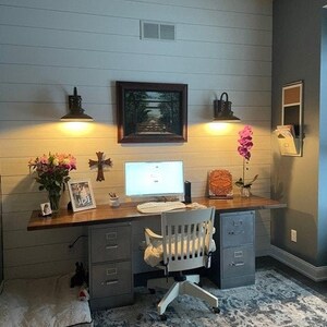 May include: A home office workspace with a white desk, two file cabinets, a chair, and a computer. The walls are white with wood paneling and there are two black wall sconces with light bulbs. There is a framed picture above the desk and a small potted plant on the desk.