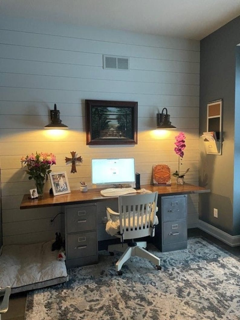 May include: A home office workspace with a white desk, two file cabinets, a chair, and a computer. The walls are white with wood paneling and there are two black wall sconces with light bulbs. There is a framed picture above the desk and a small potted plant on the desk.