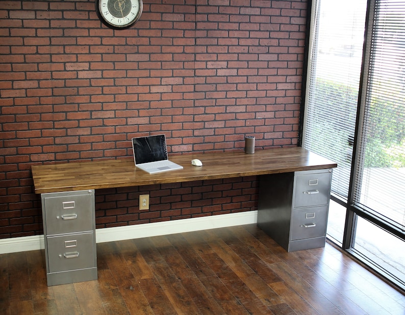 May include: A wooden desk with two metal file cabinets. The desk has a light wood finish and the file cabinets are silver. The desk is in front of a brick wall and a window.