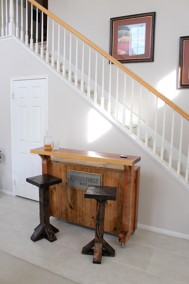 May include: A wooden bar with a metal sign that reads "Armenian Family Bar". The bar has two wooden stools with a dark brown finish. The bar is in a home with a white staircase and a framed picture on the wall.
