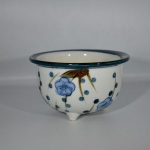 May include: A white ceramic planter with a blue and brown floral design. The planter has a dark blue rim and three small feet.