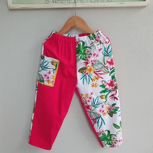May include: Trousers featuring a split design with one solid hot pink leg and one white leg patterned with colourful tropical flowers and green leaves. The trousers include an elastic waistband and a single patch pocket on the pink side.