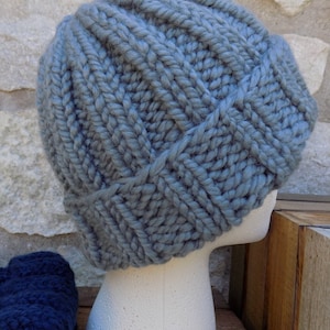May include: A chunky knit beanie in a soft, muted blue-gray color. The hat features a ribbed design and is displayed on a white mannequin head. The thick yarn creates a textured appearance, perfect for cold weather.