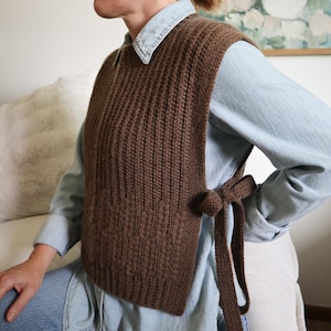 Może przedstawiać: Brązowa dzianinowa kamizelka z prążkowaną fakturą i bocznym wiązaniem. Kamizelka jest noszona na jasnoniebieskiej koszuli dżinsowej. Kamizelka ma dekoracyjny wzór na dolnej części.