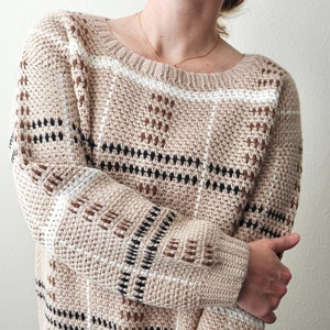 Könnte beinhalten: Ein beiger Häkelpullover mit einem karierten Muster in Braun, Schwarz und Weiß.