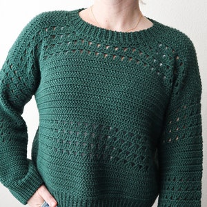 Puede incluir: Un suéter de crochet verde oscuro con un patrón de encaje en las mangas y el escote. El suéter es corto y tiene un ajuste holgado.