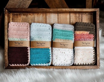 May include: Four crocheted cotton cloths in a wooden box. The cloths are in various colours and patterns, including pink, grey, teal, and brown. Each cloth has a brown label that reads "The Cozy Kast Cotton Cloths".