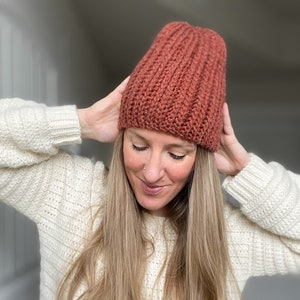 May include: A woman wearing a rust-colored crocheted beanie hat. The hat has a ribbed texture and is pulled down over her forehead.