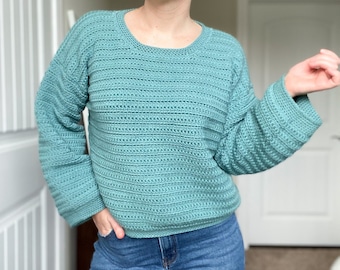 May include: A teal blue crocheted sweater with a round neckline and long sleeves. The sweater is made with a textured stitch and has a relaxed fit.