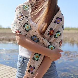 Può includere: Uno scialle in crochet beige con accenti floreali colorati. Lo scialle è drappeggiato sulle spalle di una persona ed è realizzato con un motivo a quadrati della nonna.