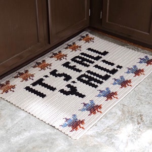 Puede incluir: Una alfombra de bienvenida de crochet blanco con texto negro que dice "It's Fall Y'all" y un borde de hojas de crochet de colores.