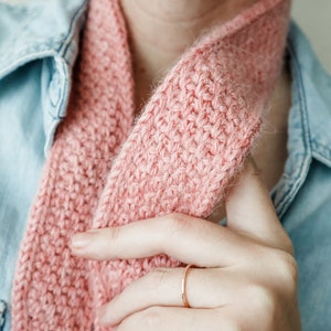 K&ouml;nnte beinhalten: Nahaufnahme einer rosafarbenen Strickm&uuml;tze, die um den Hals einer Person getragen wird, die ein Jeanshemd tr&auml;gt. Die M&uuml;tze wird von der Hand der Person gehalten, die einen d&uuml;nnen goldenen Ring tr&auml;gt.