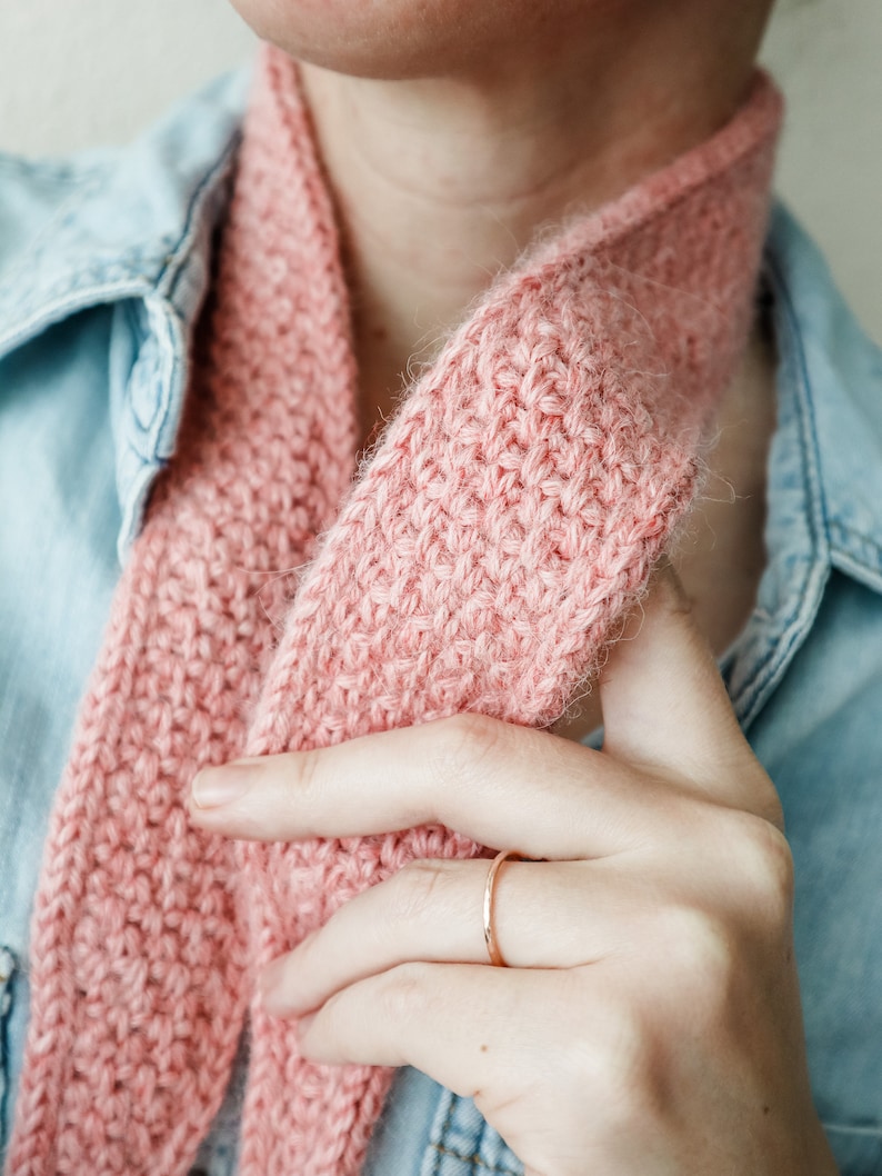 K&ouml;nnte beinhalten: Nahaufnahme einer rosafarbenen Strickm&uuml;tze, die um den Hals einer Person getragen wird, die ein Jeanshemd tr&auml;gt. Die M&uuml;tze wird von der Hand der Person gehalten, die einen d&uuml;nnen goldenen Ring tr&auml;gt.
