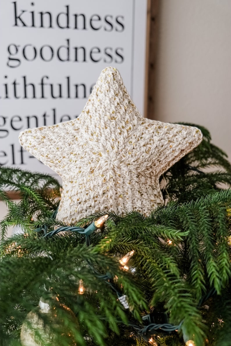 May include: A white crocheted star with gold glitter accents sits atop a green Christmas tree with white lights.