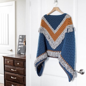 Puede incluir: Un poncho de crochet azul con diseño de cuello en V. El poncho presenta un patrón de chevron en dorado y gris, con flecos en la parte inferior y las mangas. Cuelga de una percha de madera contra una puerta blanca.