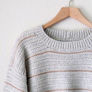 May include: A grey and beige striped crocheted jumper with a round neckline, hanging on a wooden hanger.