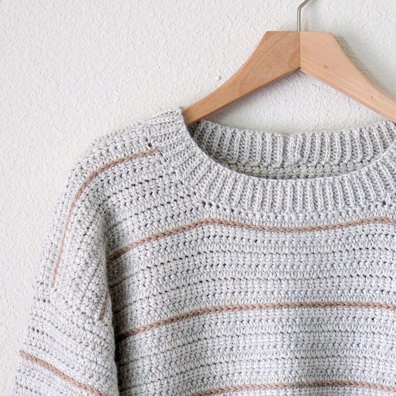 May include: A grey and beige striped crocheted jumper with a round neckline, hanging on a wooden hanger.