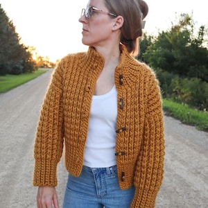 Può includere: Cardigan lavorato a maglia grossa color giallo senape con collo alto e chiusure a toggle. Il maglione corto ha maniche lunghe e viene indossato sopra una canotta bianca e jeans blu. È un capo fatto a mano e confortevole.