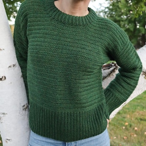 May include: A forest green crochet sweater with a ribbed hem and cuffs. The sweater has a crew neck and long sleeves. The model is wearing blue jeans. The sweater is made of a soft, textured yarn.