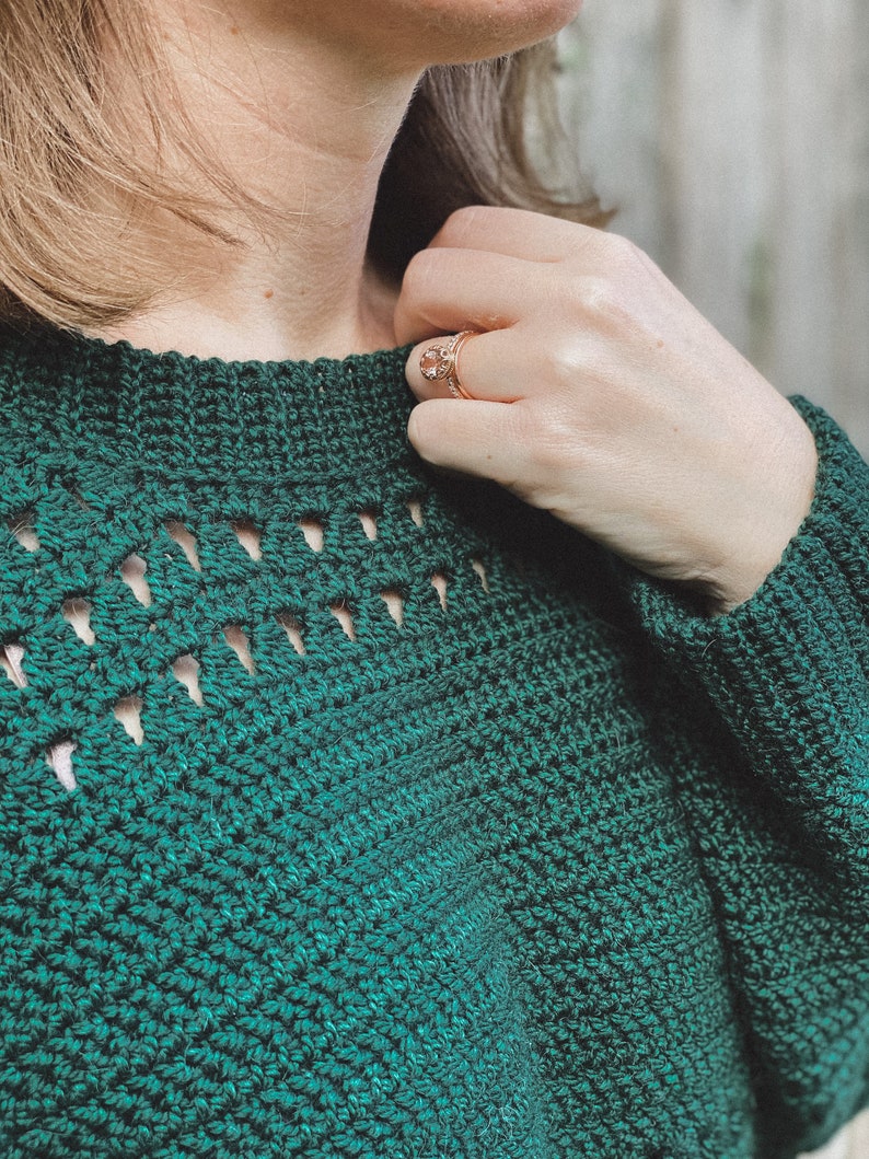 May include: A close-up of a green crocheted sweater with a delicate openwork pattern around the neckline.