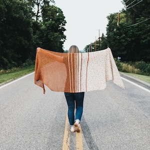 May include: A person wearing a brown and white striped crochet shawl with a brown and white striped pattern. The shawl is draped over the person's shoulders and is held in place by their hands. The person is standing on a paved road with trees in the background.