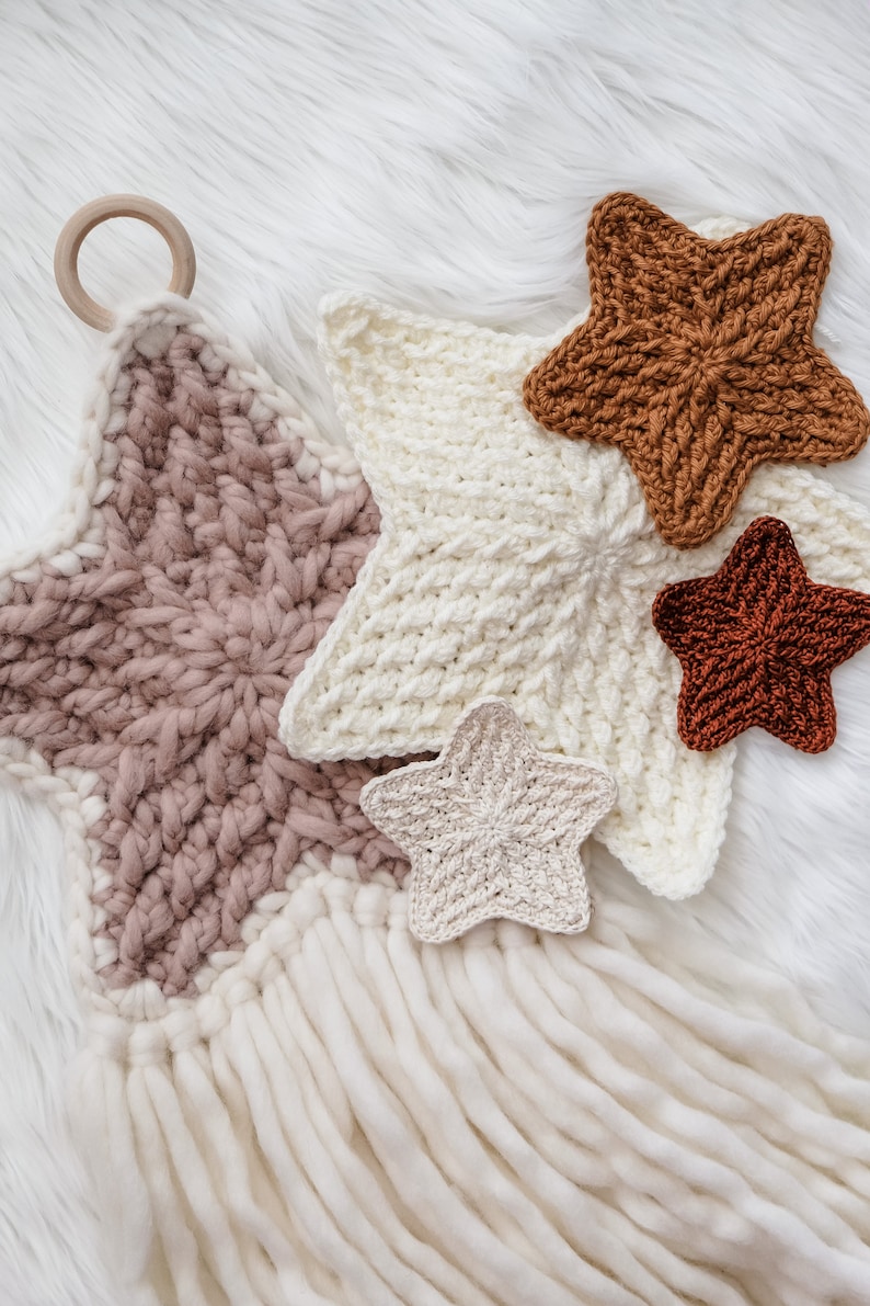 May include: A set of five crocheted stars in various shades of brown, pink, and white. The stars are hanging from a wooden ring and have long white yarn tassels.