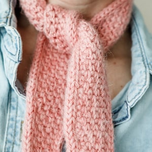 K&ouml;nnte beinhalten: Ein rosa gestrickter Schal, der um den Hals einer Person gebunden ist. Der Schal ist aus weichem, flauschigem Garn gefertigt und hat ein schlichtes, elegantes Design.
