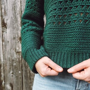 May include: A green crocheted sweater with a geometric pattern. The sweater is shown being held by a person wearing blue jeans.