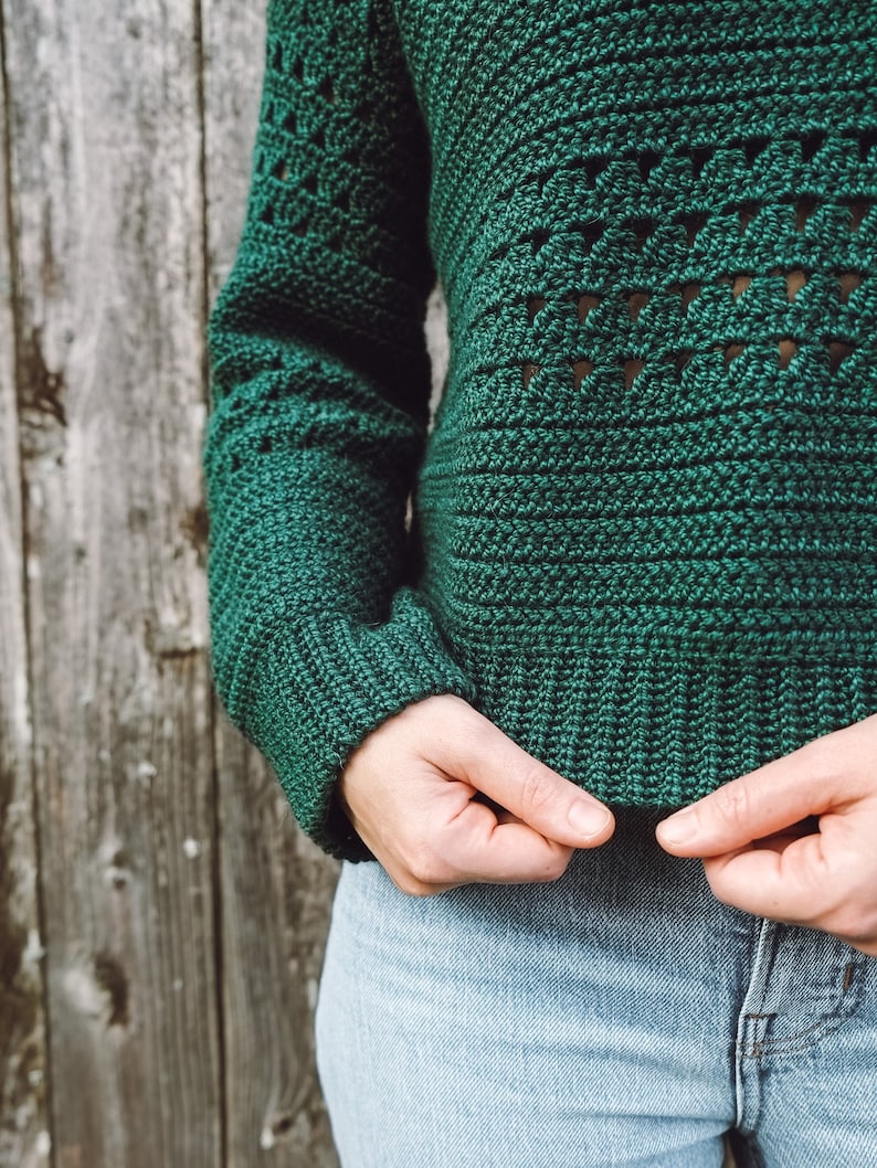 May include: A green crocheted sweater with a geometric pattern. The sweater is shown being held by a person wearing blue jeans.