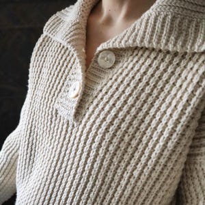 May include: A cream-colored knitted sweater with a wide collar and two button closures.