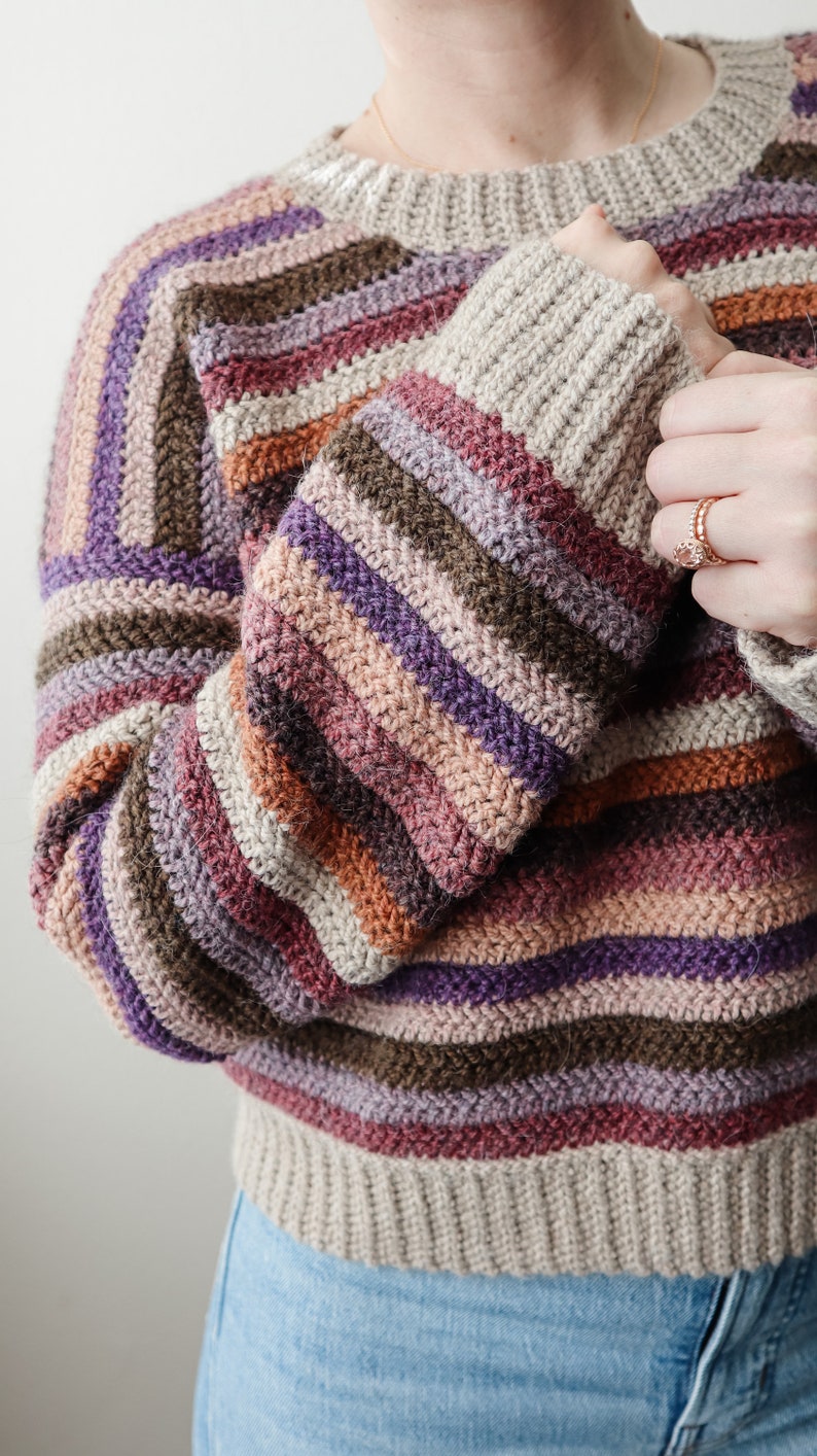 Pu&ograve; includere: Un maglione a righe in crochet con una variet&agrave; di colori, tra cui viola, marrone, rosa e beige. Il maglione ha un collo rotondo e maniche lunghe.