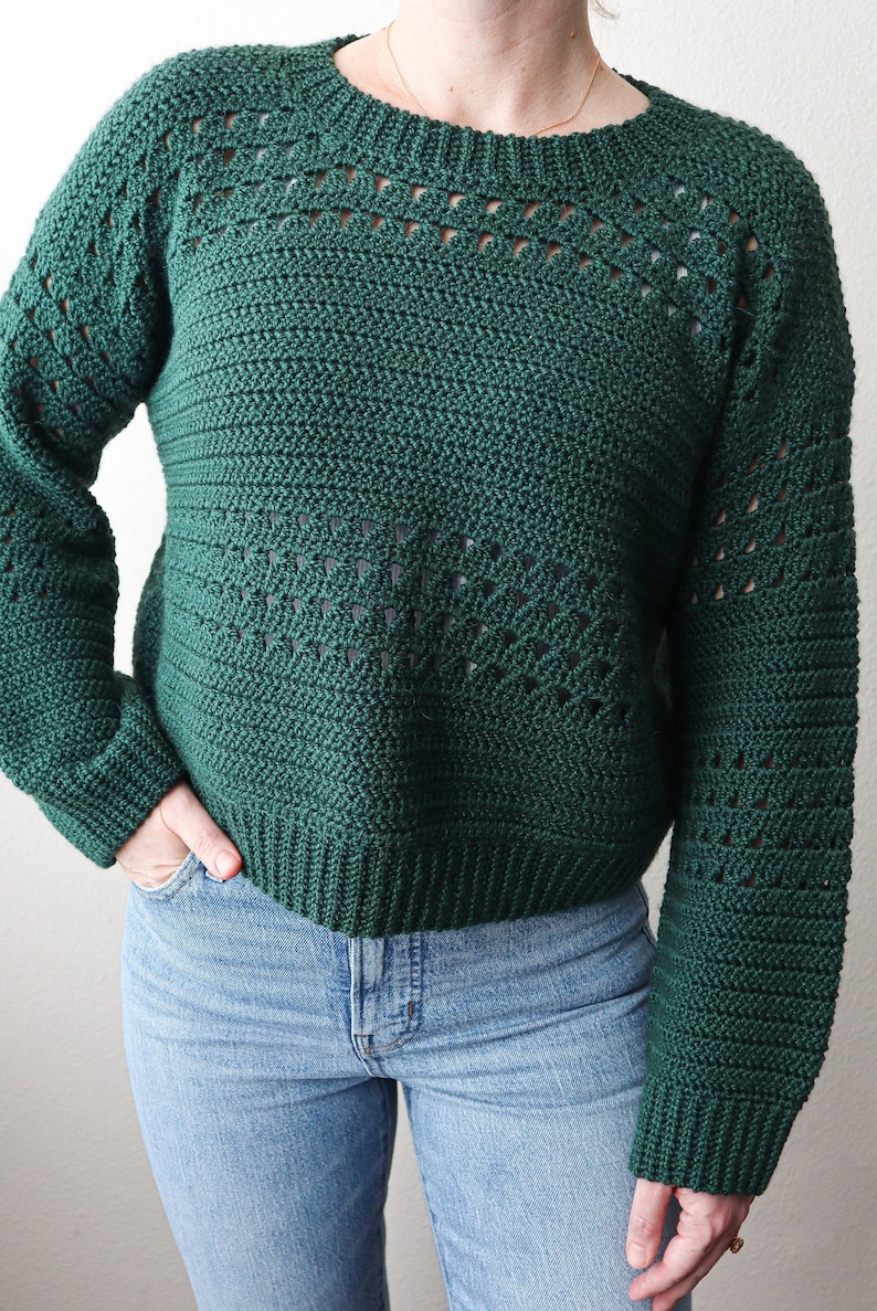 May include: A dark green crocheted sweater with a lacy pattern on the sleeves and neckline. The sweater is cropped and has a relaxed fit.