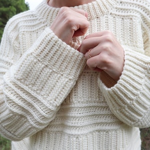 K&ouml;nnte beinhalten: Ein wei&szlig;er H&auml;kelpullover mit gerippter Struktur und langen &Auml;rmeln. Der Pullover hat eine lockere Passform und einen runden Ausschnitt.