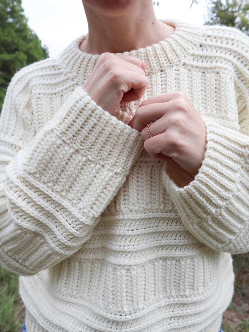K&ouml;nnte beinhalten: Ein wei&szlig;er H&auml;kelpullover mit gerippter Struktur und langen &Auml;rmeln. Der Pullover hat eine lockere Passform und einen runden Ausschnitt.