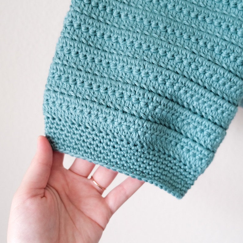 May include: A close-up of a teal crocheted fabric with a textured pattern. The fabric is held in a hand.