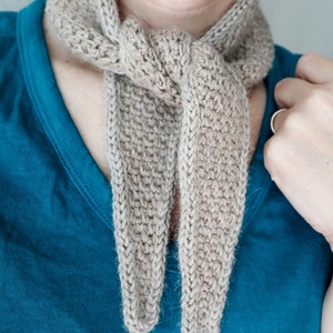K&ouml;nnte beinhalten: Ein beigefarbener H&auml;kel-Schal, der um den Hals einer Person gebunden ist. Der Schal ist aus weichem, flauschigem Garn gefertigt und hat ein schlichtes, elegantes Design.